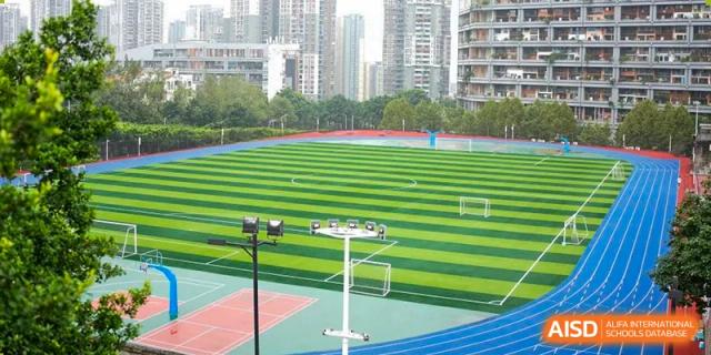 The neatly maintained football field and running track of CHONGQING WEIMING SCHOOL, surrounded by city buildings.