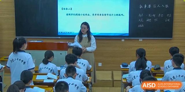 A female teacher from Dalian No.24 High School Cambridge International A-Level Centre stands in front of a digital screen, presenting a lesson to students seated at their desks.