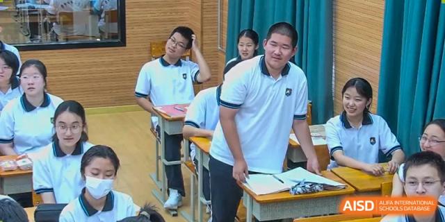 A male student and a male teacher from Dalian No.24 High School Cambridge International A-Level Centre smile in a classroom while standing between student desks.