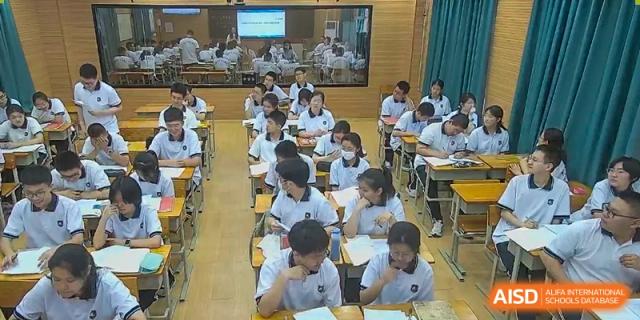 A class of students from Dalian No.24 High School Cambridge International A-Level Centre, all wearing white shirts, focus on their work at their desks in a brightly lit classroom.