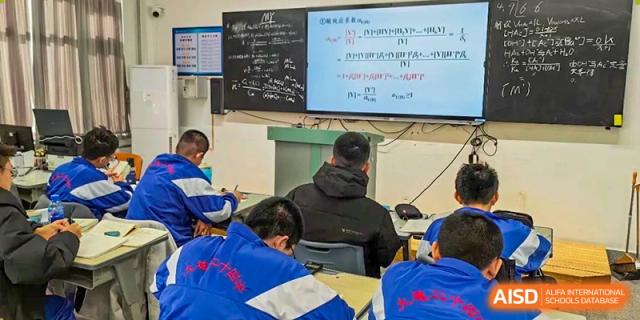 Students from Dalian No.24 High School Cambridge International A-Level Centre in blue uniforms work at their desks in a classroom, with mathematical equations visible on the whiteboard and an illuminated screen.