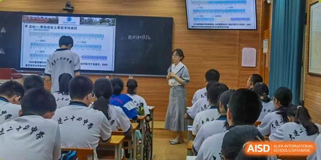 A female teacher from Dalian No.24 High School Cambridge International A-Level Centre addresses a classroom full of students, standing near a blackboard.