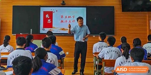A male teacher from Dalian No.24 High School Cambridge International A-Level Centre stands in front of a projector screen, actively teaching a class. The screen displays Chinese characters.