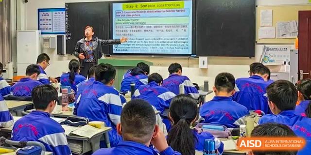 Students from Dalian No.24 High School Cambridge International A-Level Centre in blue uniforms are seated at their desks, watching a teacher present information on a screen.