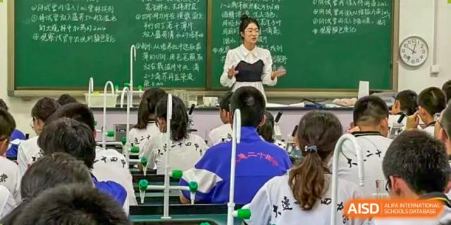 A female teacher from Dalian No.24 High School Cambridge International A-Level Centre stands in front of a green blackboard, teaching students who are seated in what appears to be a laboratory or specialized classroom.