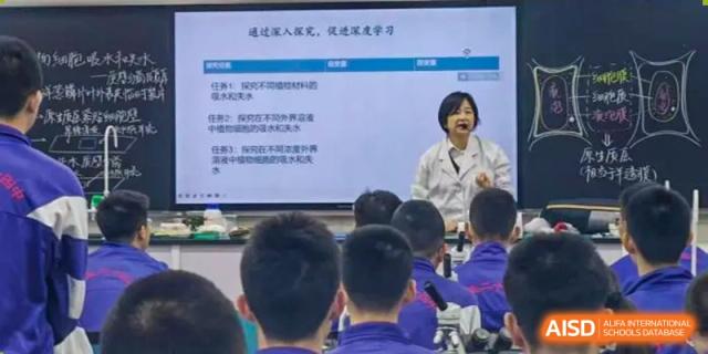 A classroom at Dalian No.24 High School Cambridge International A-Level Centre where students face a large screen displaying a lesson, with a teacher visible on the screen.
