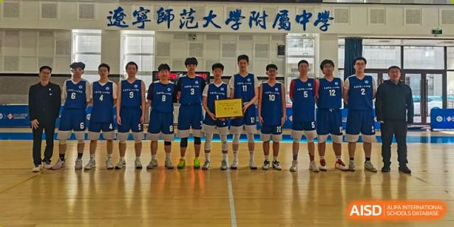 A boys' indoor volleyball or basketball team from Dalian No.24 High School Cambridge International A-Level Centre poses together in a gymnasium, all wearing matching blue and white uniforms and holding a trophy or award.