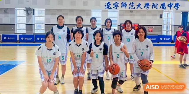 A girls' basketball team from Dalian No.24 High School Cambridge International A-Level Centre poses on a court, wearing white and black uniforms. One of the girls holds a basketball.