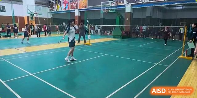 Two students from Dalian No.24 High School Cambridge International A-Level Centre play a game of badminton on an indoor court with a green surface and net.
