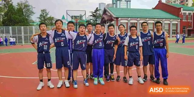 An outdoor group photo of a boys' basketball team from Dalian No.24 High School Cambridge International A-Level Centre, with the students in dark blue jerseys smiling and standing in front of an outdoor court and school buildings.
