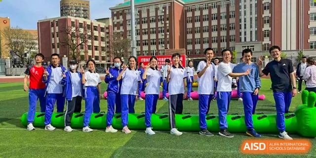 A group of students from Dalian No.24 High School Cambridge International A-Level Centre, dressed in white and blue uniforms, stand outside next to several bright green objects.
