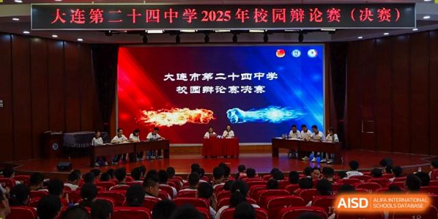 An interior view of an assembly or graduation ceremony at Dalian No.24 High School Cambridge International A-Level Centre, showing a large screen with event text and an audience seated below.