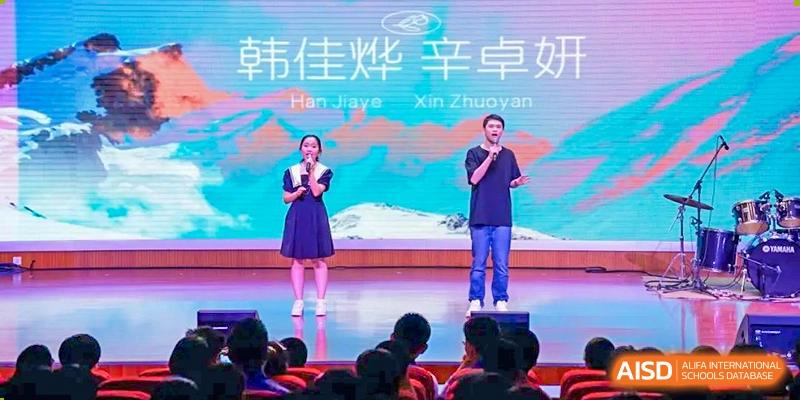 Two speakers or performers from Dalian No.24 High School Cambridge International A-Level Centre stand on a stage with a banner overhead displaying Chinese characters and English names.