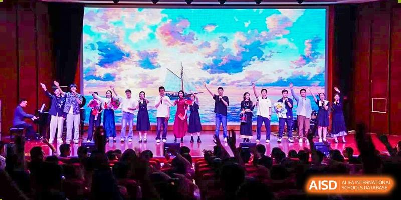 Students from Dalian No.24 High School Cambridge International A-Level Centre celebrate and perform on a stage illuminated by bright lights and a colorful sky backdrop in front of a cheering audience.