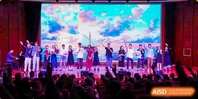 Students from Dalian No.24 High School Cambridge International A-Level Centre celebrate and perform on a stage illuminated by bright lights and a colorful sky backdrop in front of a cheering audience.