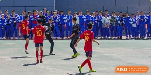 Students from Dalian No.24 High School Cambridge International A-Level Centre play soccer/football on a grass field, with one player in a red jersey kicking the ball past a blue-jersey opponent.