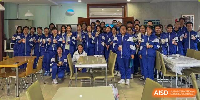 A large group of students from Dalian No.24 High School Cambridge International A-Level Centre, all wearing blue tracksuits, poses for a cheerful group photo indoors.