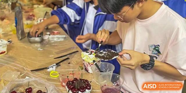 Two students from Dalian No.24 High School Cambridge International A-Level Centre participate in a hands-on activity, possibly baking or decorating food, involving a small cake with cherries.