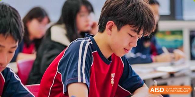 A male student at Dulwich College Suzhou is in class, wearing a red and blue sports jersey with a school crest. He is looking down at his desk.