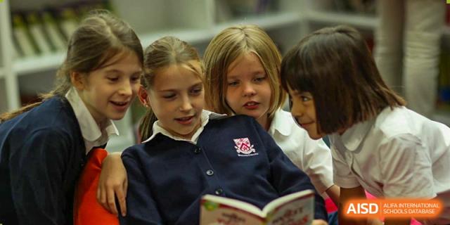 Two young female students at Dulwich College Suzhou are sitting together and looking at a book with smiles on their faces. They are in a classroom.