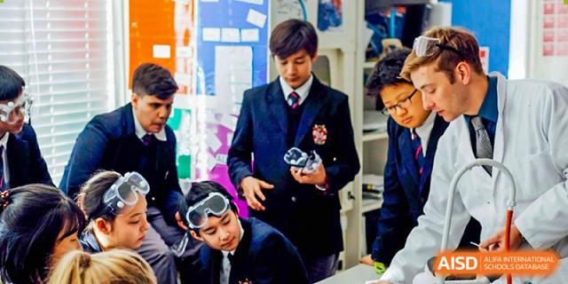 Two male students from Dulwich College Suzhou are in a classroom wearing a school uniform and holding a small object, while another student is on the floor wearing safety goggles.