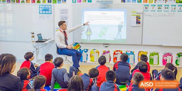A male teacher at Dulwich College Suzhou is sitting in front of a whiteboard with a group of young students. He is pointing at a graphic on the board.