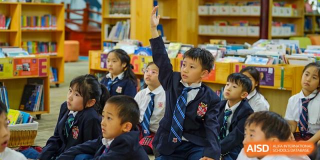 A group of young students at Dulwich College Suzhou, wearing school uniforms, are seated in a classroom with one student raising his hand to answer a question.