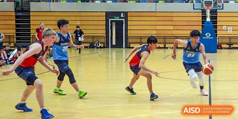 Two male students from Dulwich College Suzhou are playing a sport in a basketball gymnasium, with one student in a red and blue sports jersey running on a court.
