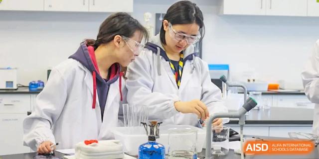 Two students from Dulwich International Suzhou High School Programme conducting a chemistry experiment in a lab, wearing lab coats and safety goggles.