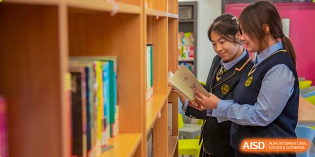 A Grand International School student in uniform selecting or reading a book from a shelf in the school library.