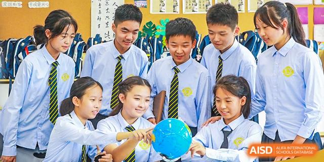 A group of Grand International School students in uniform gathered around a table, looking down at a small blue globe or model during a class activity.