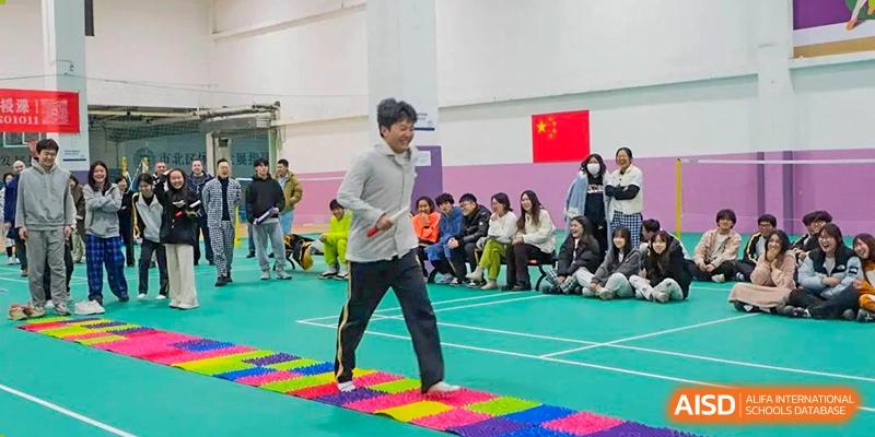 A male participant from Grand International School walking across a colorful reflexology mat or textured path in an indoor gymnasium, with spectators watching.