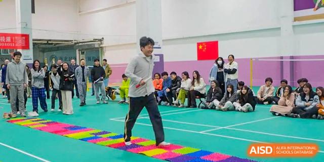 A male participant from Grand International School walking across a colorful reflexology mat or textured path in an indoor gymnasium, with spectators watching.