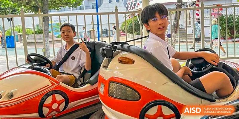 Two Grand International School students in uniform sitting in a carnival-style ride (bumper car or similar) and smiling.