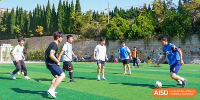 Grand International School students playing a game of soccer or football on a green outdoor field under a sunny sky.