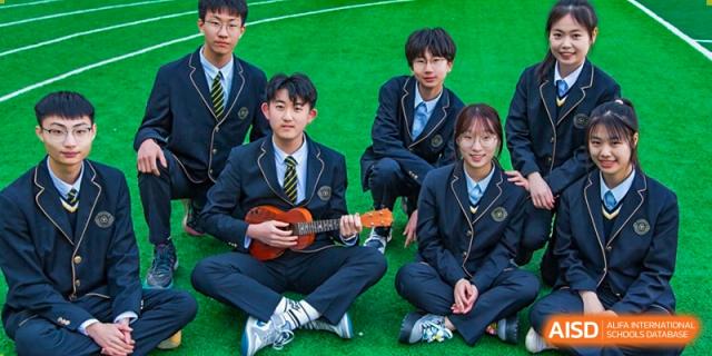 A small group of Grand International School students in uniform sitting together outdoors, with one student holding a ukulele.