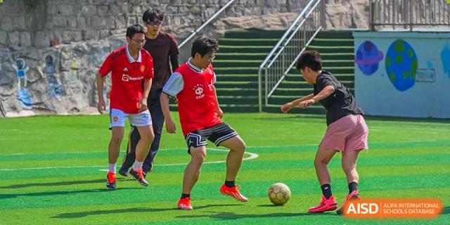 Grand International School students playing an intense game of soccer on a paved or outdoor court near stairs.