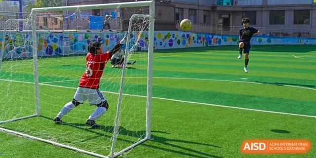 A Grand International School student, possibly a goalkeeper, jumping to defend the soccer goal on an artificial turf field.