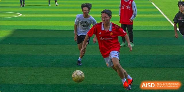 A Grand International School student in a red jersey dribbling a soccer ball during a fast-paced game on a green field.