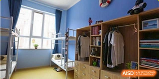 An interior shot of a student dormitory room at Grand International School, featuring wooden storage cabinets, clothing, a bookshelf, and a metal bunk bed near a window.