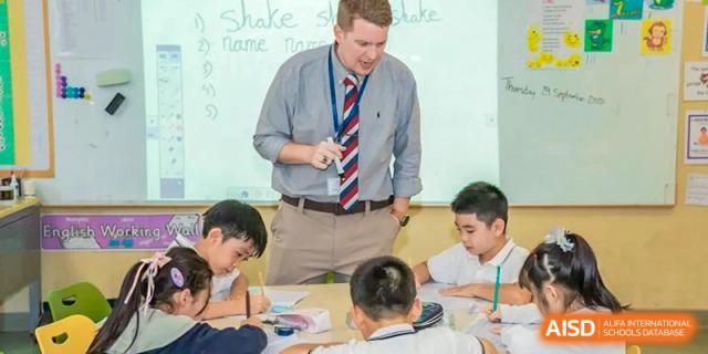 A teacher standing at the front of a classroom, talking to a group of students seated at a table at HD Qingdao School.