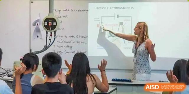 A teacher pointing to a whiteboard with a diagram and text about electromagnetism, with students looking on at HD Qingdao School.