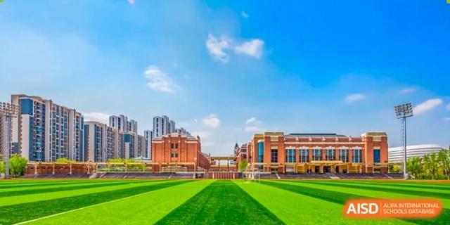 A large red brick building with many windows and a clock tower, surrounded by a green lawn and blue sky with clouds at HD Qingdao School.