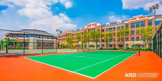 A green and orange sports court with a white net, surrounded by trees and a red brick building at HD Qingdao School.