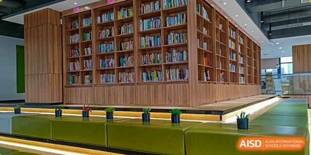 A section of the library at Hongyi Cambridge International School (HYCIS) with wooden bookshelves filled with books and a low, green seating area.