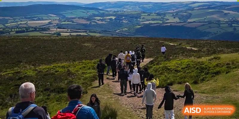Students from Hongyi Cambridge International School (HYCIS) on a group hike on a grassy hill, following a dirt path with a wide green landscape in the background.