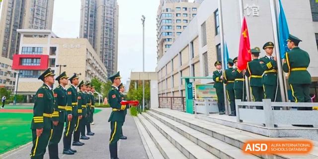A person in a military or security uniform standing near the steps and modern buildings of the Hongyi Cambridge International School (HYCIS) campus entrance.