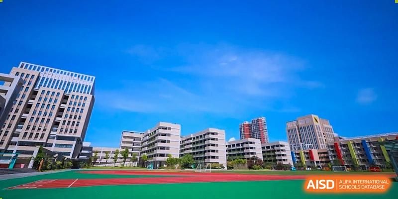 The modern, white academic buildings of Hongyi Cambridge International School (HYCIS)viewed across a green and red sports field under a clear blue sky.