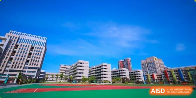The modern, white academic buildings of Hongyi Cambridge International School (HYCIS)viewed across a green and red sports field under a clear blue sky.