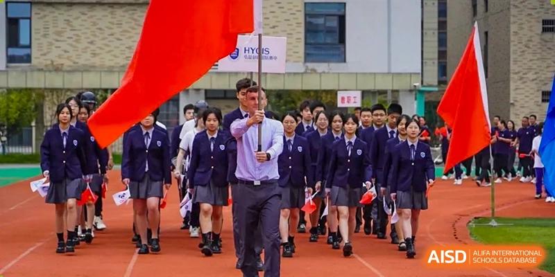 A student carrying a large red flag leads a procession of students in navy blue uniforms at Hongyi Cambridge International School (HYCIS).